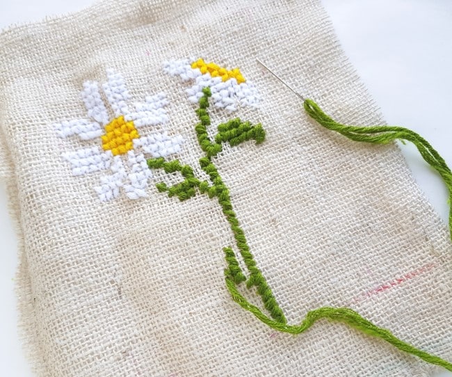 Easy Cross Stitch Pattern - two daisies stitches on burlap fabric, with the green stem and leaves nearly done being stitched, needle with green yarn inside